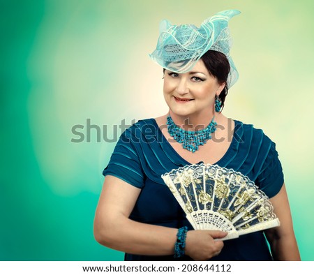 Old woman posing in blue dress, blue organza hat, turquoise accessories and holding fan in hand