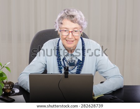 Senior grey haired teacher looking at laptop screen. She is teaching on-line English (or any) language. Teacher is wearing blue blouse and dark blue stone necklace with pendant.