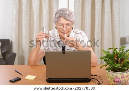 Senior glasses woman is taking pill during reading bad news on the Internet. Grey haired retired woman is sitting at desk in front of laptop monitor with glass of water.