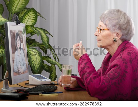 Old woman taking a pill during medical consultation on-line. Grey haired woman in crimson cape sitting opposite monitor. On the screen doctor is consulting her. Side shot