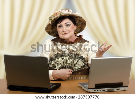 Skillful acting coach sits in front of two laptops and works with learners on-line. Senior actress is wearing a dress of the 19th century and tapestry hat. She holds retro guipure fan in right hand
