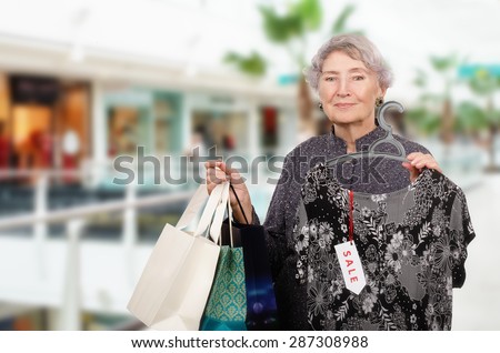 Amazing old woman shopping in the mall with three shopping bags in left hand. In right hand, she holds elegant long dress on hangers with tag sale. There is mall out of focus in the background