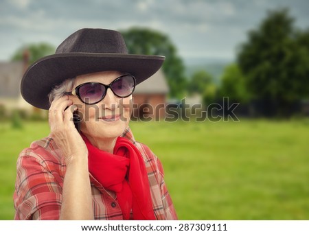 Smiling aged cowgirl is talking on smart phone. Old woman in sunglasses is wearing black cowboy hat and red plaid shirt. Old cowgirl in red neckerchief is posing on countryside background out of focus