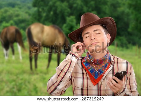 Young cowboy in earphones is listening country music with smart phone. He is wearing brown cowboy hat, plaid shirt and colored bandana neckerchief