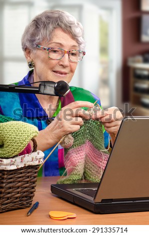 Aged woman is knitting in front of laptop. She is member of on-line knitting club. Retired woman knows the mobility and dexterity required for knitting helps improve brain function.