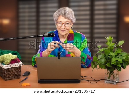 Senior woman is knitting in front of laptop. She is teaching on-line learners the art of knitting. Experienced teacher is using two web-cams to help students in training.
