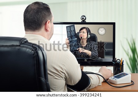 Middle-aged man measures his blood pressure in front of virtual doctor. In the meantime, telemedicine physician is carefully looking at his brain x ray picture in the monitor. Horizontal shot