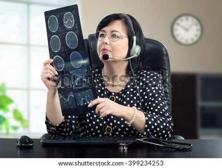 Telemedicine doctor in headphones is looking at x ray picture of brain carefully. Black haired doctor in glasses sitting at black desk. She is wearing a polka dot blouse. Horizontal shot