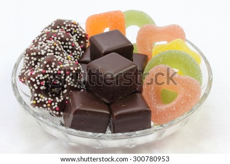 Various Christmas goodies like dominoes, jelly fruits and fudge stars on a glass plate on white background
