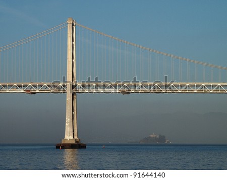 Sun shines on the Bay Bridge with Alcatraz island in the distance with a lite layer of fog.