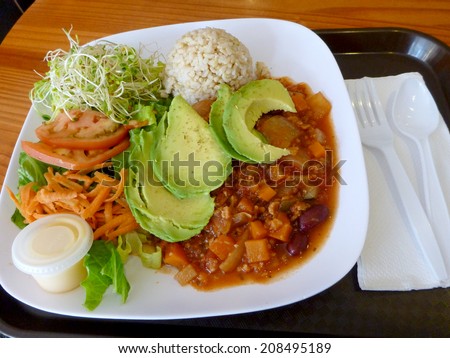 Totally Vegan Special - Veggie Chili, Avocado, Brown rice and Salad on a plate on a tray on a table with a napkin, plastic spoon, and fork
