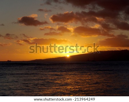 dramatic lighting as Sunsets behind Waianae mountains with light reflecting on ocean and illuminating the sky with boats sailing on the water off Waikiki on Oahu, Hawaii.