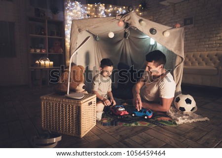 Father and son are playing with toy cars on carpet road in blanket fort at night at home.