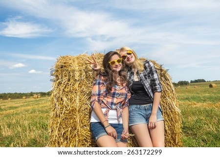 Student girls in checkered shirts and denim hot pants are resting on large round straw bale at big farm harvested cereal field background and navy blue sky. Young girls in sunglasses and show V sign