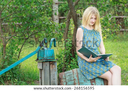 Blonde student girl in summer garden is reading opened book with blue cover