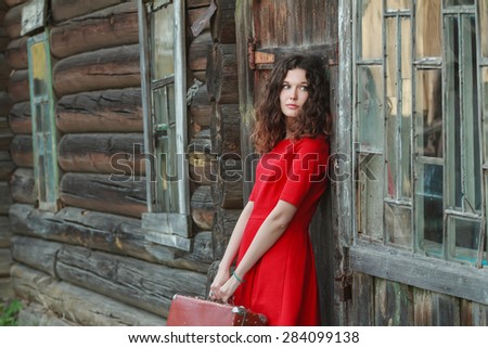 Melancholy woman is leaning on wooden wall of old log cabin with retro suitcase