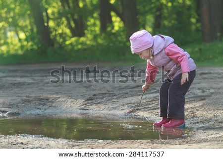 Toddler preschooler girl is exploring summer puddle wearing red gums thin tree twig in hand