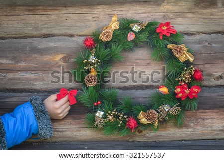 Human hand is holding red bow and hanging on rustic log cabin wall with Christmas wreath