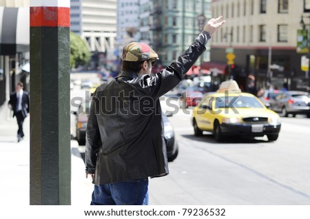 A man wearing a funky cap and black leather jacket at the side of the street hailing a taxi cab