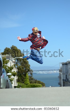A man wearing a funky cap, tartan Jacket, blue jeans and boots  jumps up high in the street