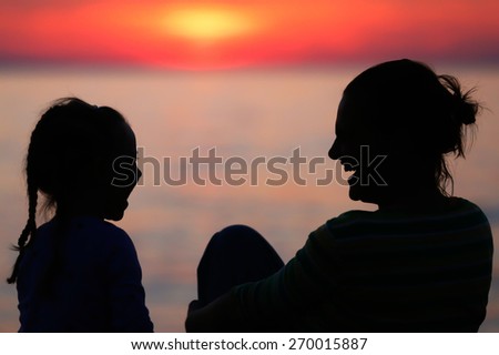 Silhouette of a mother and daughter laughing and sharing a tender bonding moment on the beach for Mothers day at colorful sunset.