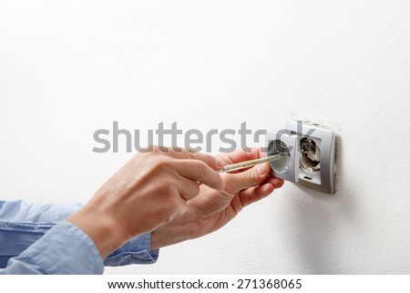 Electrician installing a silver wall-mounted AC power socket with a screwdriver on a white wall, renovating home.