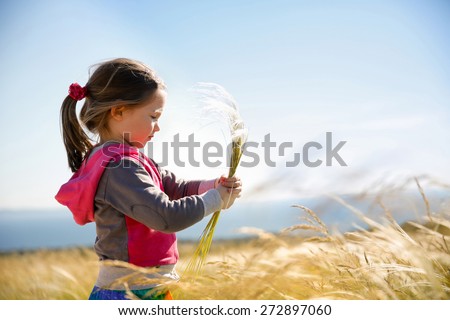 Cute little girl picking and gathering brown grasses on a meadow with the wind blowing through her long hair and sea in the background. Allergy, hay fever and hypersensitivity concept.