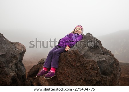 Cheerful, cute little girl resting on a volcanic boulder of Mount Etna, volcano on the island of Sicily, Italy. Active family lifestyle, traveling family concept.