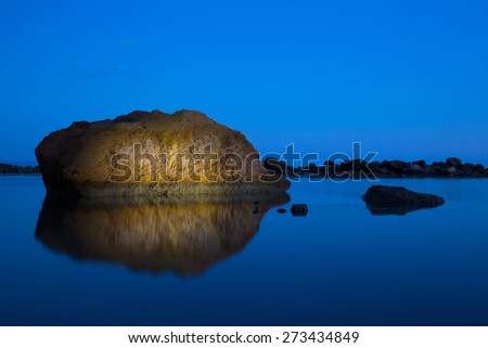 Uniquely lit, dramatic rock formation in a tranquil sea, reflecting on the surface of deep blue colored water. Calmness, tranquility, meditation, calm before the storm concept, blue background.