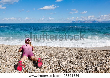 Girl lying on her belly, observing sea and enjoying free time on the beach, dressed in wetsuit and a hat for sun protection. Family and children on vacation, summer fun concept.