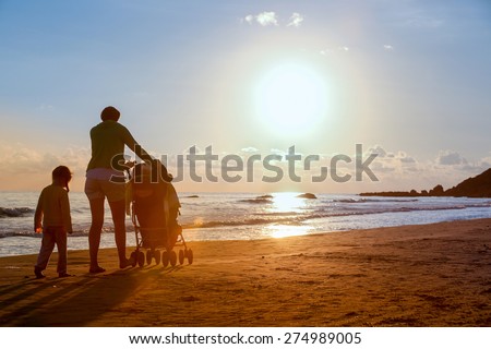 Silhouette of mother walking with her daughter and baby, pushing a stroller on a sandy beach in late summer, enjoying the evening chill. Family vacation, traveling with children concept.