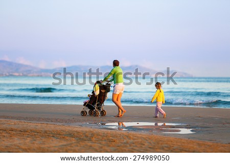 Single mother walking in silence with her daughter and baby, pushing a stroller on a sandy beach in late summer, enjoying the evening chill. Family vacation, traveling with children concept.