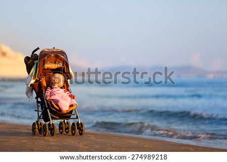 Baby in a stroller on a sandy beach, peacefully looking into infinity and sea waves with great interest, enjoying late summer evening by the sea. Family vacation, traveling with children concept.