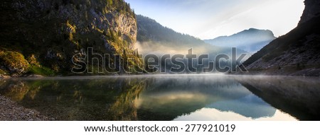 Extraordinary, fairy tale, mythological mountain lake scenery in a glacial valley, with sun shining to the rocky slope and morning mist above the lake surface with perfect reflection.