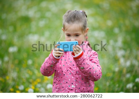 Little curious girl photographing with her smart phone, exploring nature and standing in a dandelion meadow. Active lifestyle, curiosity, pursuing a hobby, technology and kids concept.