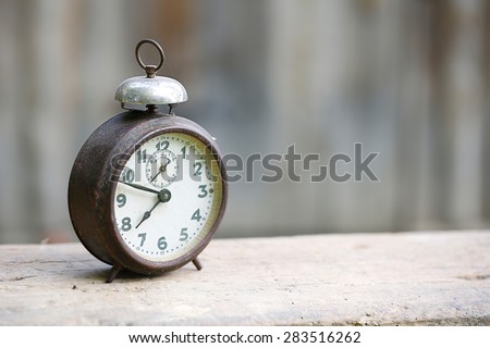 Vintage metal analog alarm clock with Arabic numbers and wind-up mechanism, sitting on a wooden bench with retro background. Time is now, time is money, old times concept.