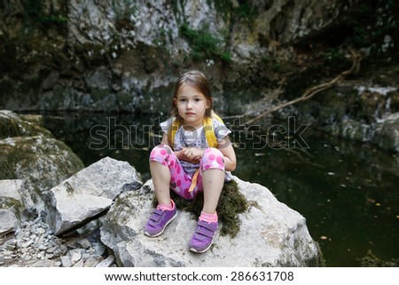 Tired but happy little girl resting on a big rock by a small pond, during a forest hike, exploring big nature. Active, healthy and natural lifestyle concept.