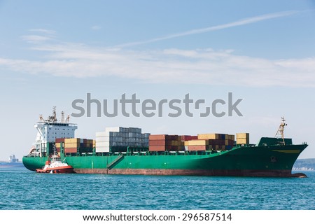 Big container ship, loaded with various colorful containers, waiting to be assisted and escorted to the port by a towboat. Global transportation, global business, consumerism concept and background.