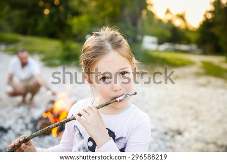 Little girl eating roasted marshmallow by a self-made campfire on family camping trip, with her dad in the background. Active natural lifestyle, fun family time concept.