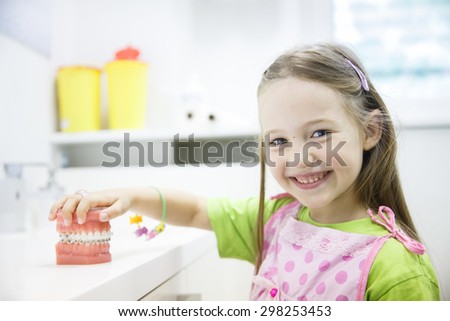 Little girl holding an artificial model of human jaw with dental braces in orthodontic office, smiling. Pediatric dentistry, aesthetic dentistry, early education and prevention concept.