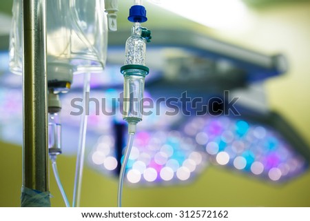 Functioning IV drip hanging on a pole in hospital, with LED surgical lights in the background. Patient, illness, treatment, hospital, medicine and healthcare abstract and concept.