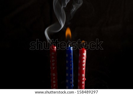 Two red and one blue birthday candles, two candles are lit, third is blown out with smoke trail, black background