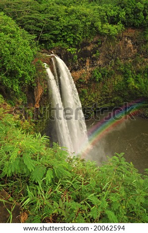 Waterfalls drop in a water pool in a lush green forest, with a rainbow rising from the mist