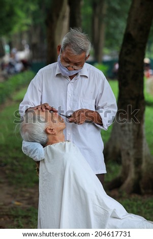 HANOI, VIETNAM - JUL 13, 2014: Unidentified old hairdresser serving an old man at a park in Hanoi capital. Many Vietnamese like to get hair cut outdoor, with cheap pay.