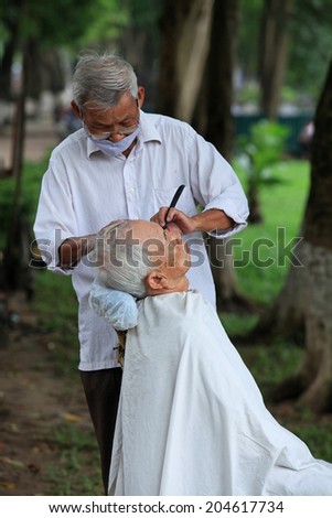 HANOI, VIETNAM - JUL 13, 2014: Unidentified old hairdresser serving an old man at a park in Hanoi capital. Many Vietnamese like to get hair cut outdoor, with cheap pay.
