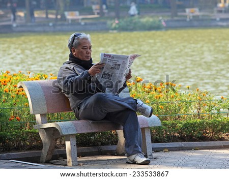HANOI, VIETNAM - JAN 22, 2014: Unidentified Vietnamese old man reading newspaper on the bank of Hoan Kiem (Sword) lake in the sunlight. Hoan Kiem lake is known as the center of Hanoi capital.