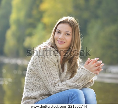Beautiful girl enjoying the autumn weather sitting outdoors with a lovely smile on her face in a warm polo-neck sweater in front of a tranquil lake