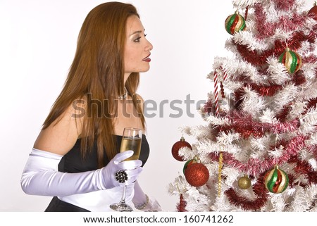 A Hispanic woman in an evening gown and white gloves, holding a glass of champagne, while looking at a decorated Christmas tree.