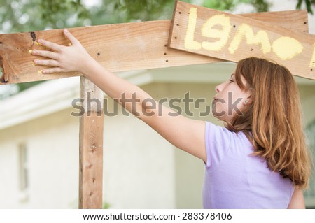 Young girl painting lemonade stand places her hand, covered in yellow paint, on the sign to make a hand print.