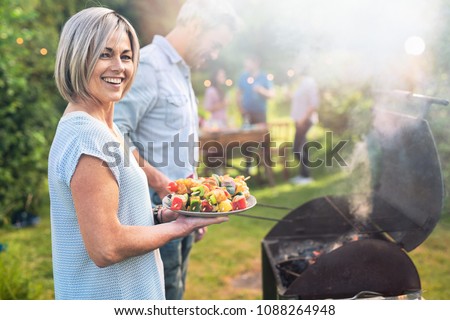 In summer. A nice couple prepares a bbq to welcome friends in the garden. She\'s holding a plate of grilled skewers